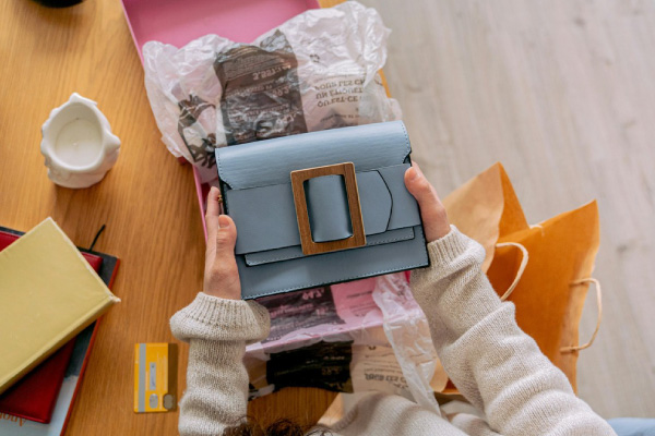 A woman holds a blue purse, resting on a table, showcasing her stylish accessory in a refined setting.